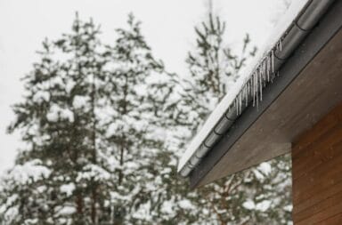 Stunning winter scene of icicles hanging from a snow-covered roof gutter with snowy pines in the background.